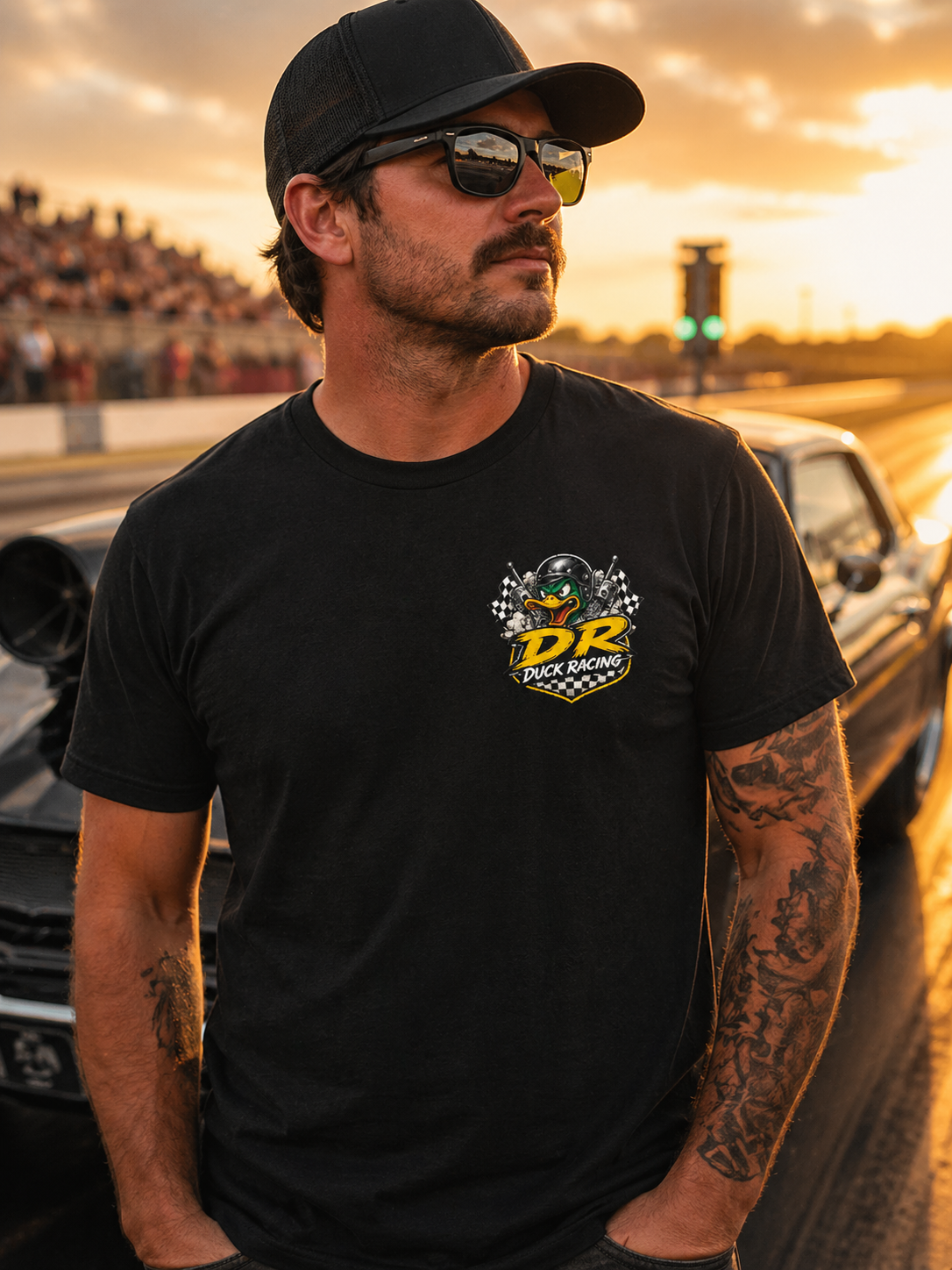 Man wearing a black t-shirt with a 'Duck Racing' logo, standing on a race track at sunset.