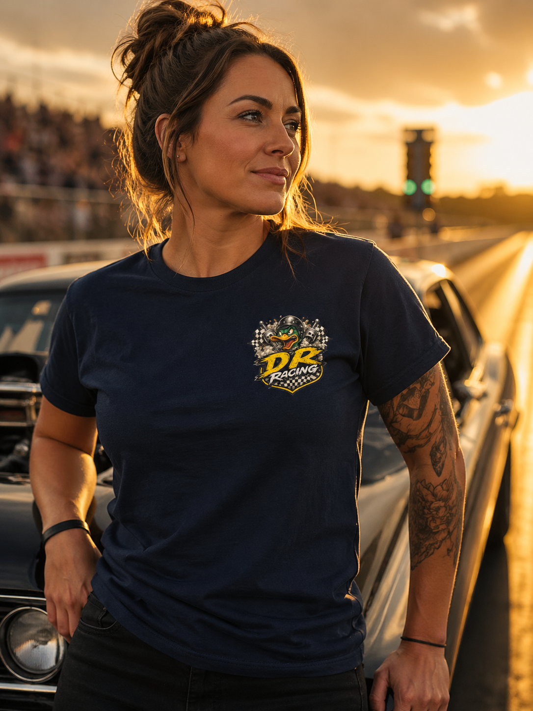 Woman wearing a navy blue t-shirt with a 'Duck Racing' logo, standing in front of cars at sunset.
