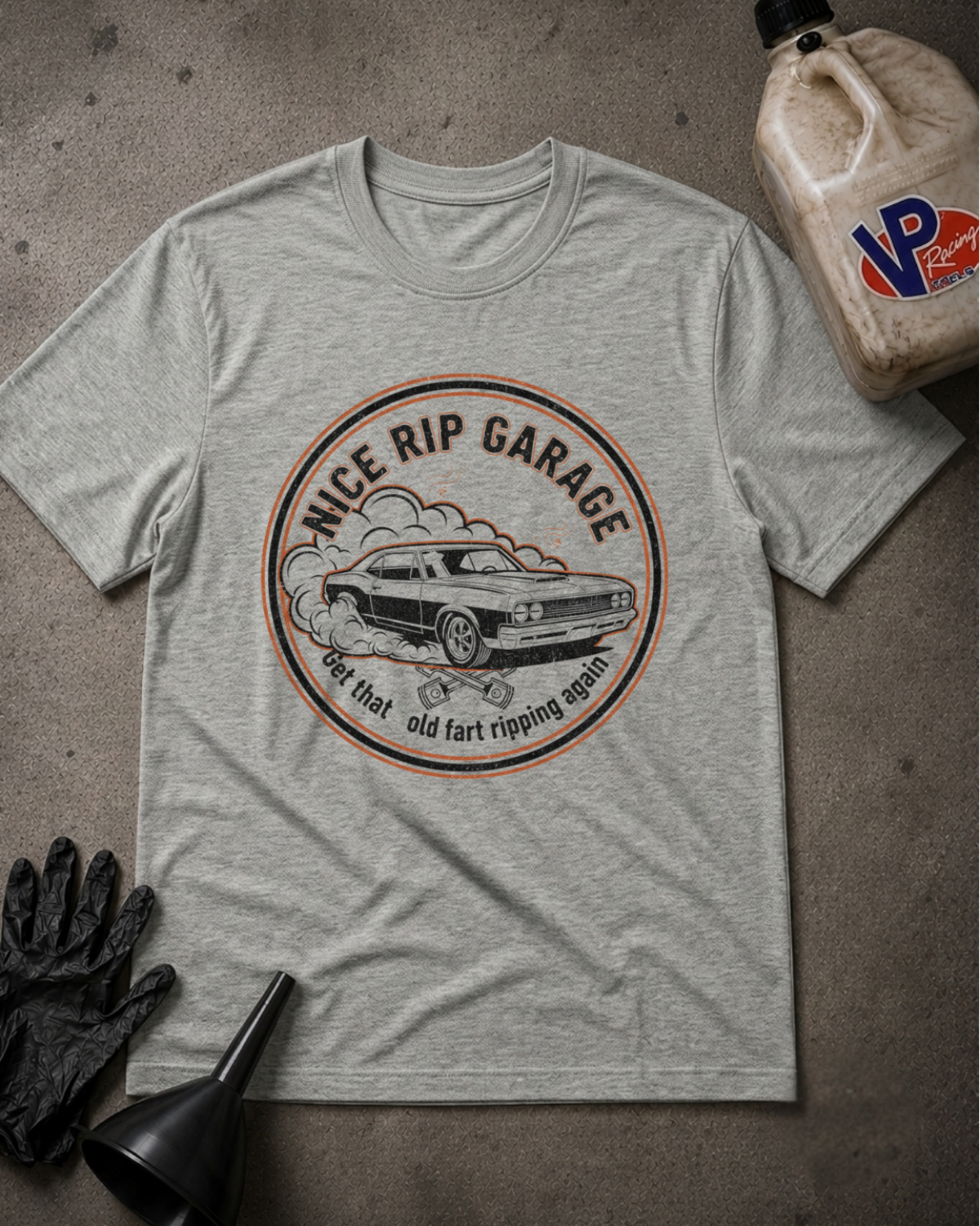 A heather gray graphic t-shirt laid flat on a textured gray surface, surrounded by automotive-themed props including a black funnel, black gloves, and a plastic jug of VP Performance oil. The t-shirt features a circular graphic with a vintage muscle car and the text 'NICE RIP GARAGE' and 'get that old fart ripping again'.
