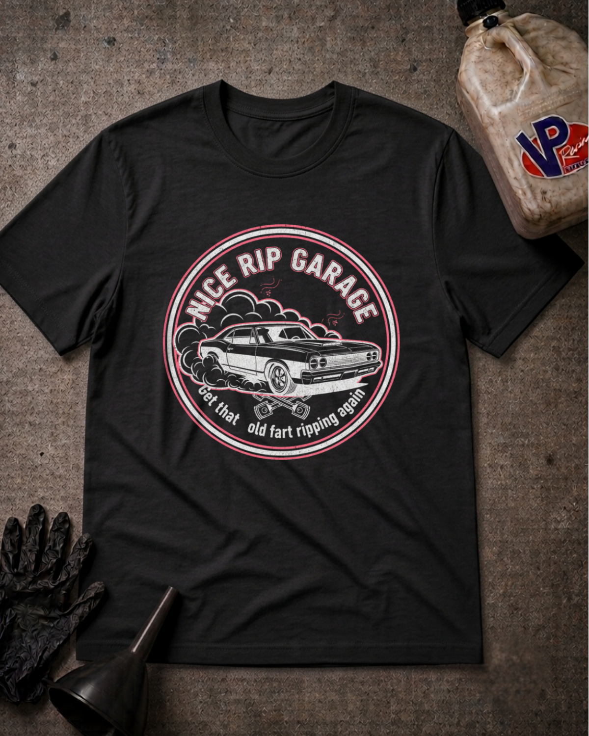 A black graphic t-shirt laid flat on a textured gray surface, surrounded by automotive-themed props including a black funnel, black gloves, and a plastic jug of VP Performance oil. The t-shirt features a circular graphic with a vintage muscle car and the text 'NICE RIP GARAGE' and 'get that old fart ripping again'.