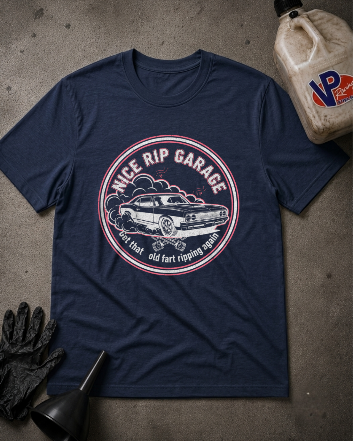 A navy blue graphic t-shirt laid flat on a textured gray concrete surface, surrounded by automotive-themed props including a black funnel, black gloves, and a plastic jug with a VP logo. The t-shirt features a circular graphic with a classic car and the text 'NICE RIP GARAGE' and 'Get that old fart ripping again'.