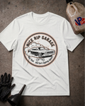 A white short-sleeve graphic T-shirt laid flat on a dark textured surface, surrounded by automotive-themed props including a black glove, funnel, and a bottle of fluid. The shirt features a circular vintage car graphic with the text 'Nice Rip Garage' and smaller text below.