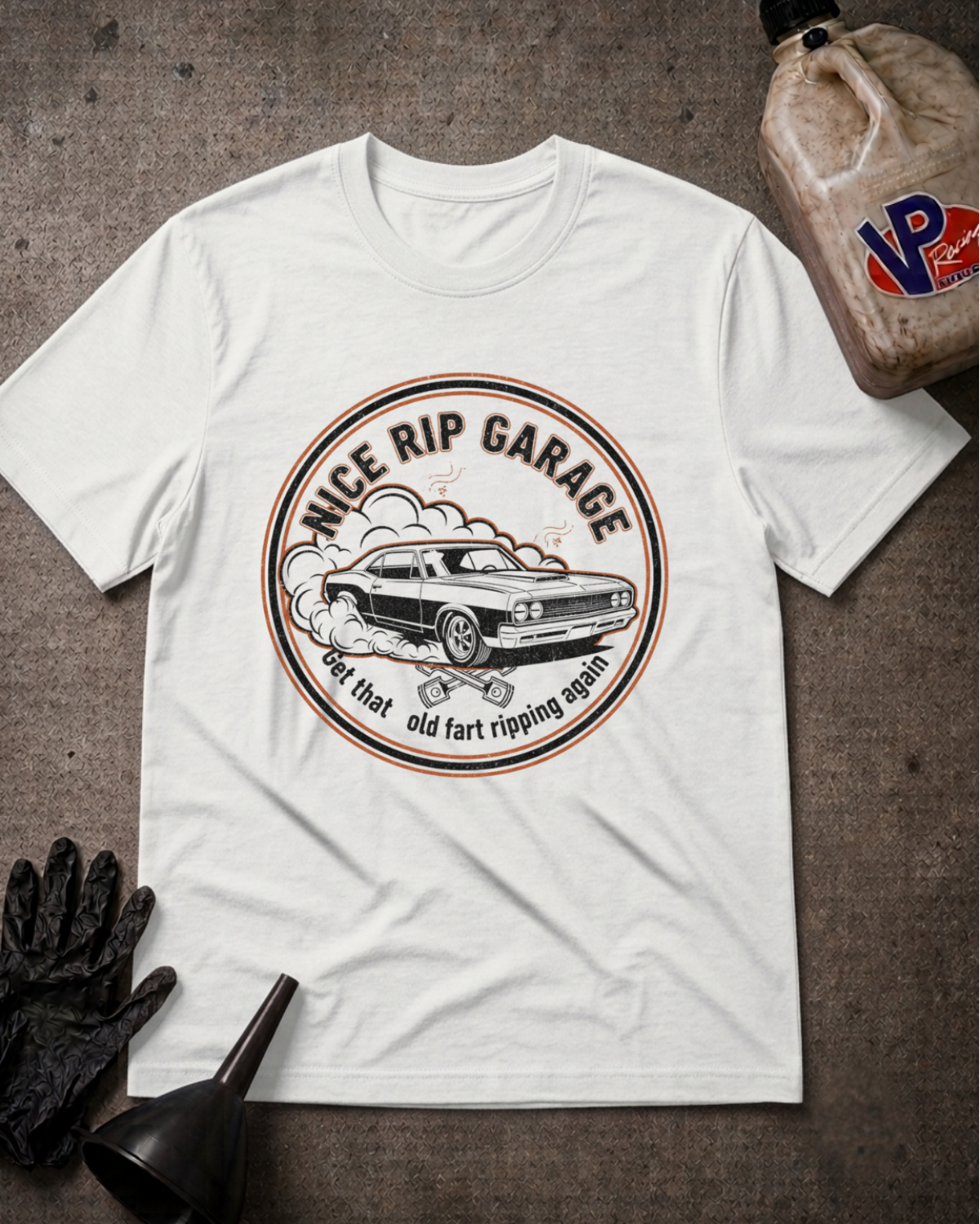 A white short-sleeve graphic T-shirt laid flat on a dark textured surface, surrounded by automotive-themed props including a black glove, funnel, and a bottle of fluid. The shirt features a circular vintage car graphic with the text 'Nice Rip Garage' and smaller text below.