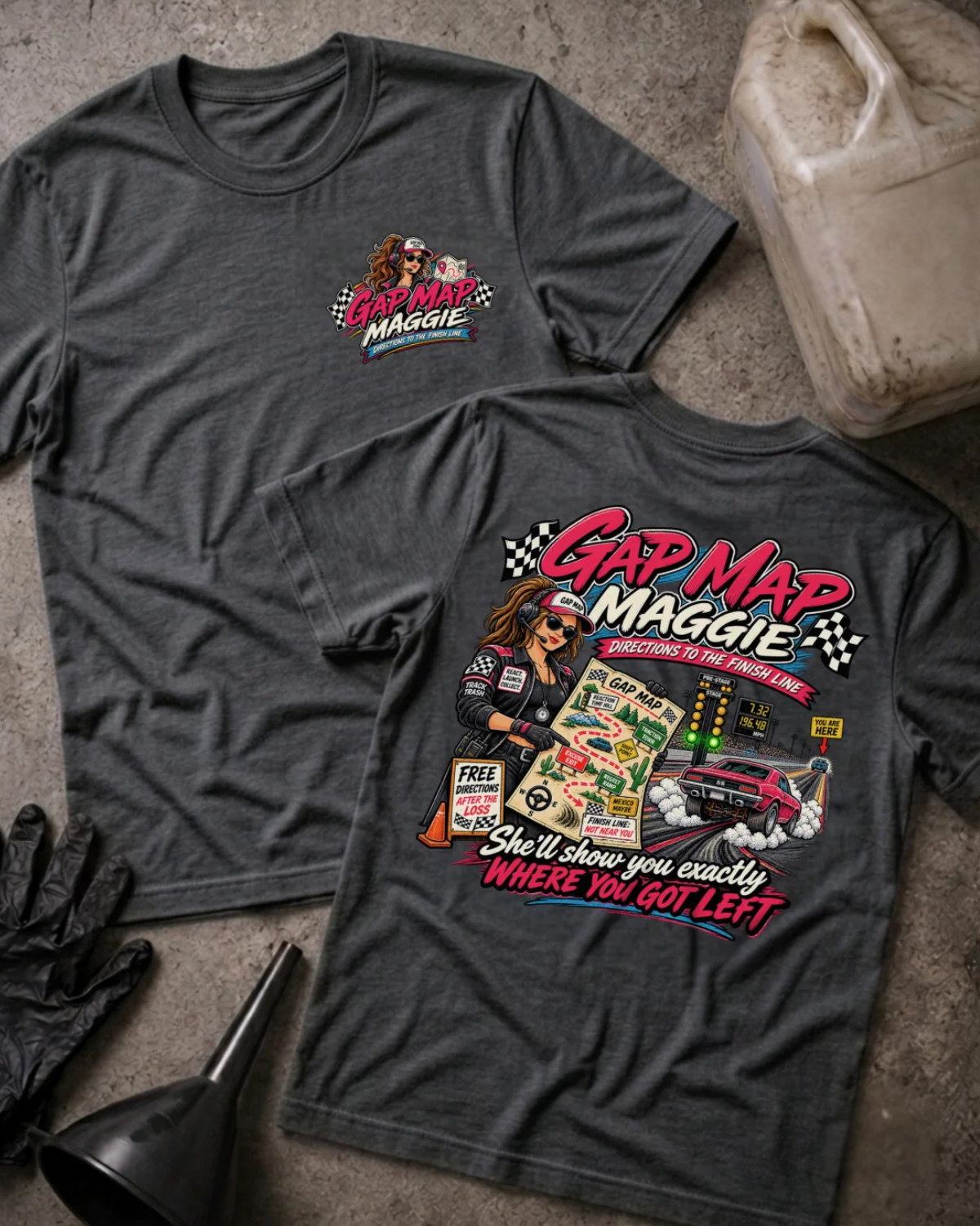 Two dark grey heather graphic T-shirts laid flat on a concrete surface, showing the front and back designs. The shirts feature a colorful 'Gap Map Maggie' racing graphic with a cartoon woman in a racing suit, checkered flags, a race car, and the phrase 'She'll show you exactly WHERE YOU GOT LEFT'.