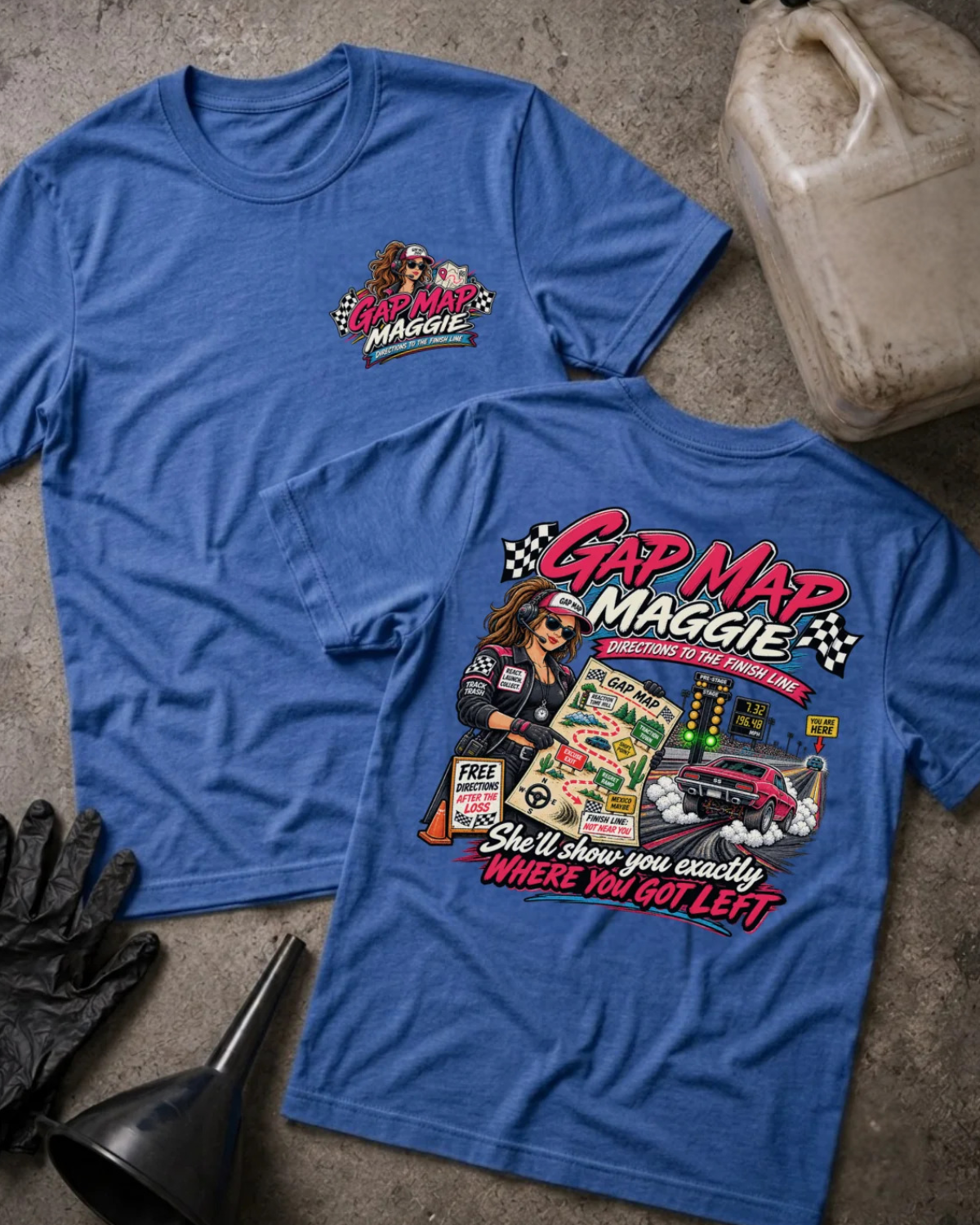 Two heather true royal graphic T-shirts laid flat on a concrete surface, showing the front and back designs. The shirts feature a colorful 'Gap Map Maggie' racing graphic with a cartoon woman in a racing suit, checkered flags, a race car, and the phrase 'She'll show you exactly WHERE YOU GOT LEFT'.