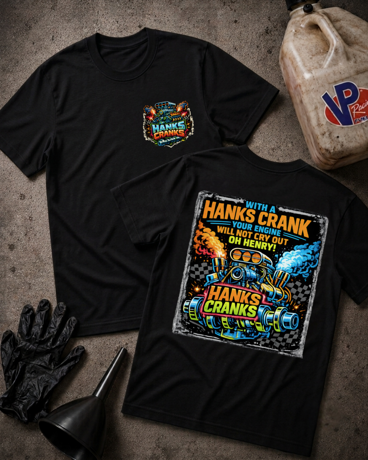 Two black graphic T-shirts laid flat on a textured gray surface, with a large colorful front chest print and a larger back graphic reading 'Hank's Crank.' Surrounding props include black gloves, a funnel, and a plastic jug with a visible VP logo.