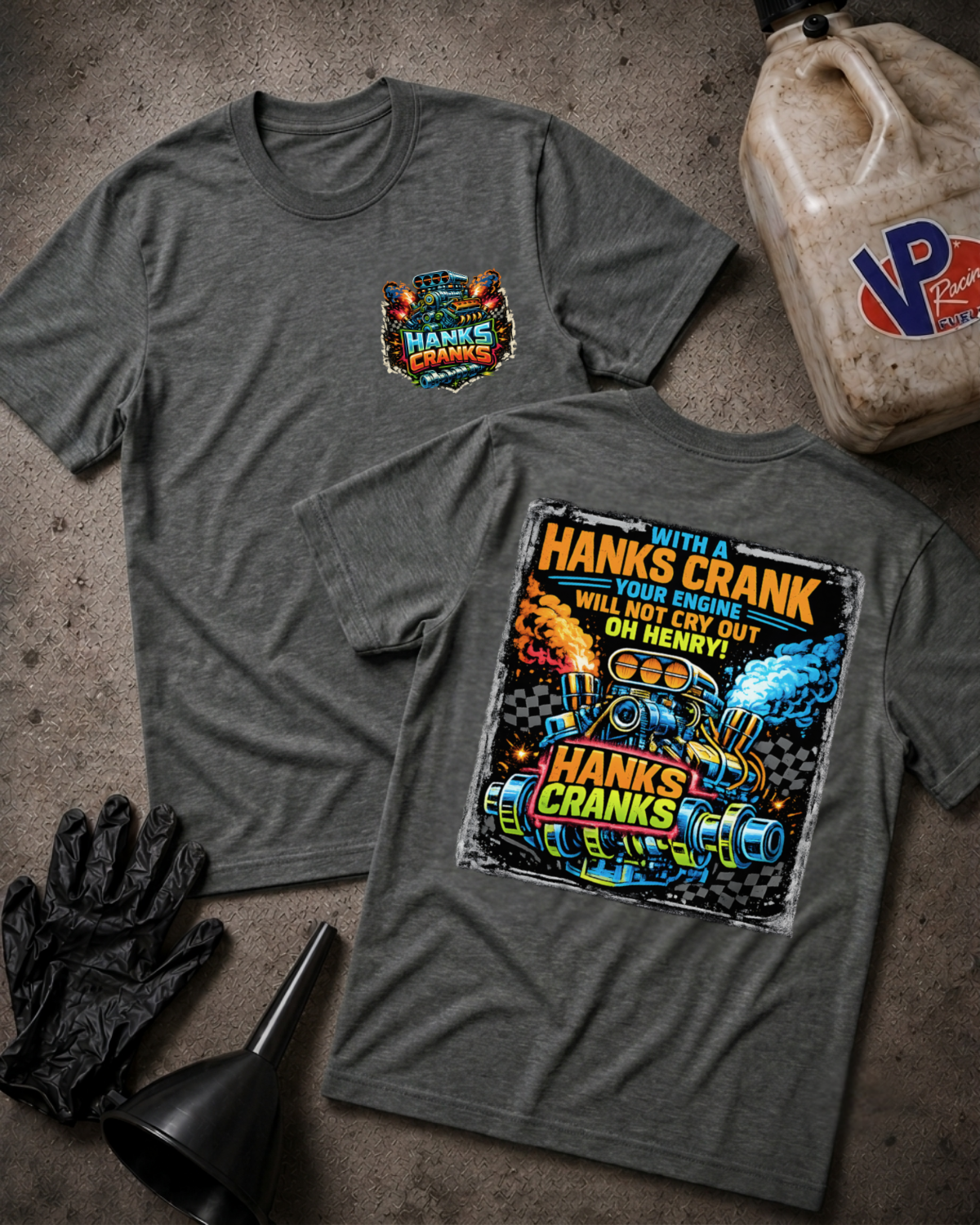 Two dark heather gray graphic T-shirts laid flat on a textured concrete surface, with a gas can, black gloves, and a black funnel as props. The front shirt shows a small multicolor chest graphic reading 'Hanks Crank', and the back shirt shows a large colorful engine graphic with the text 'WITH A HANKS CRANK YOUR ENGINE WILL NOT CRY OUT OH HENRY!'
