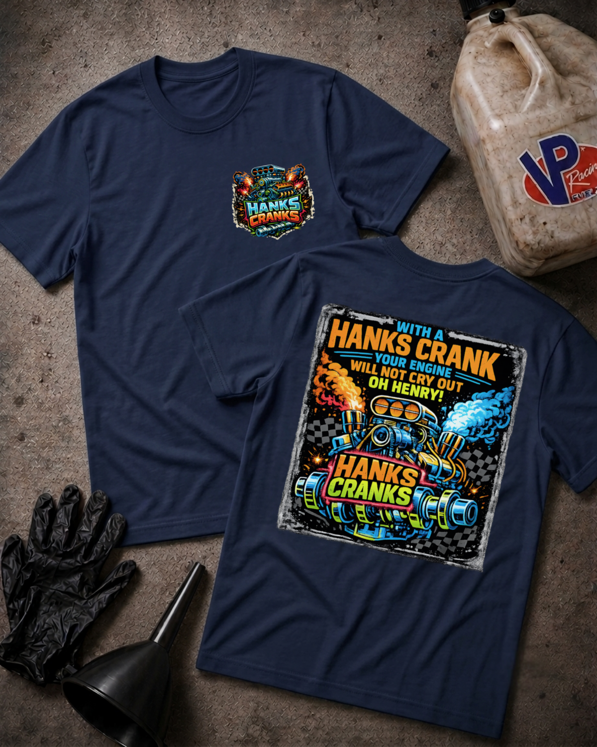 Two navy blue graphic T-shirts laid flat on a textured gray surface, with a black oil jug, black gloves, and a black funnel around them. The front shirt shows a small multicolor chest graphic reading 'Hanks Crank', and the back shirt shows a large colorful engine graphic with the text 'WITH A HANKS CRANK OUR ENGINE WILL NOT CRY OUT OH  HENRY!'.