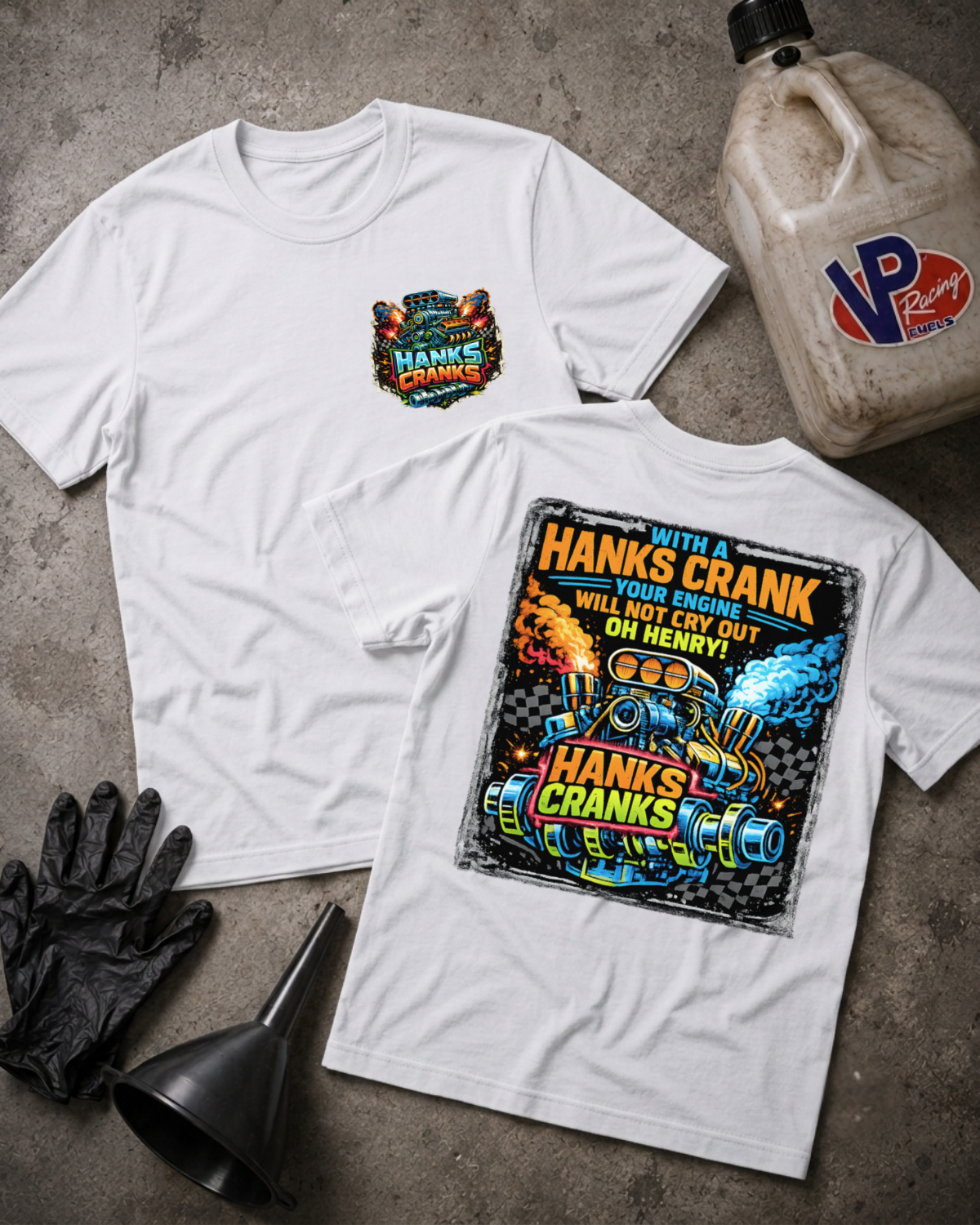 Two white graphic T-shirts laid flat on a gray concrete surface, with a black funnel, black gloves, and a VP Racing fuel jug as styled props. The front shirt shows a small multicolor chest graphic reading 'Hank's Crank,' and the back shirt shows a large multicolor engine-themed graphic with the same text.