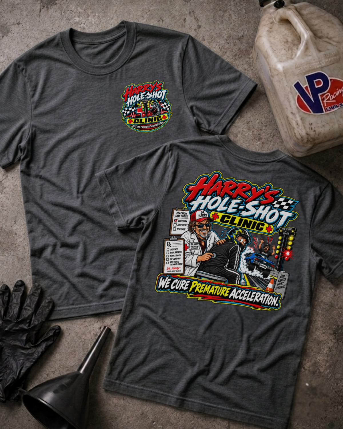 Two dark heather gray graphic T-shirts laid flat on a concrete surface, with a gas jug, gloves, and funnel as styled props. The front shirt shows a small multicolor chest graphic reading 'Harry's Hole-Shot Clinic'; the back shirt shows a large colorful racing clinic graphic with the text 'Harry's Hole-Shot Clinic' and 'We Cure Premature Acceleration.
