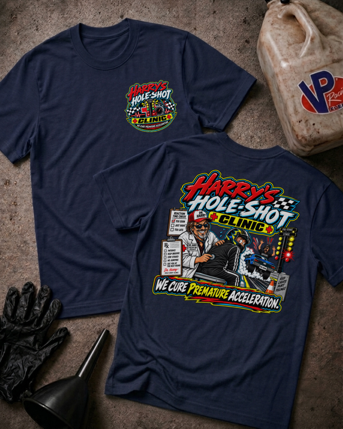 Two navy graphic T-shirts laid flat on a concrete surface, with a gas jug, gloves, and funnel as styled props. The front shirt shows a small multicolor chest graphic reading 'Harry's Hole-Shot Clinic'; the back shirt shows a large colorful racing clinic graphic with the text 'Harry's Hole-Shot Clinic' and 'We Cure Premature Acceleration.