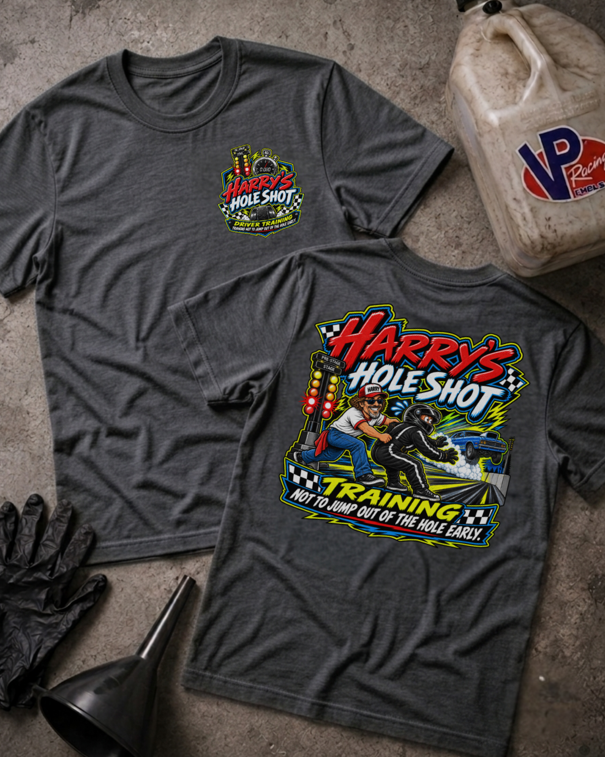 Two dark heather gray graphic T-shirts laid flat on a concrete surface, with a black funnel, black gloves, and a plastic fuel jug used as styling props. The front shirt shows a small multicolor chest graphic reading 'Harry's Hole Shot,' and the back shirt shows a large multicolor racing-style graphic with the same text.