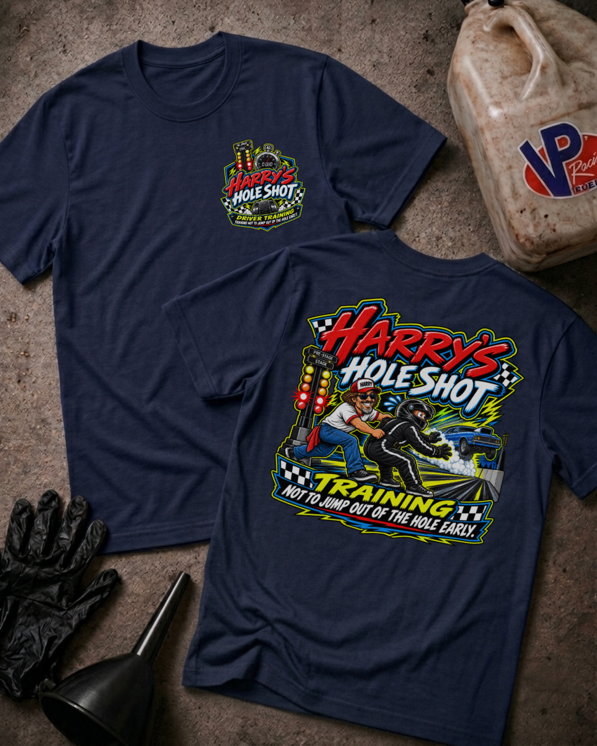 Two navy graphic T-shirts displayed on a concrete surface, with a black oil jug, black gloves, and a funnel as props. The front shirt shows a small multicolor chest graphic reading 'Harry's Hole Shot,' and the back shirt shows a large colorful racing-style graphic with the text 'Harry's Hole Shot' and 'Training Not to Jump Out of the Hole Early.