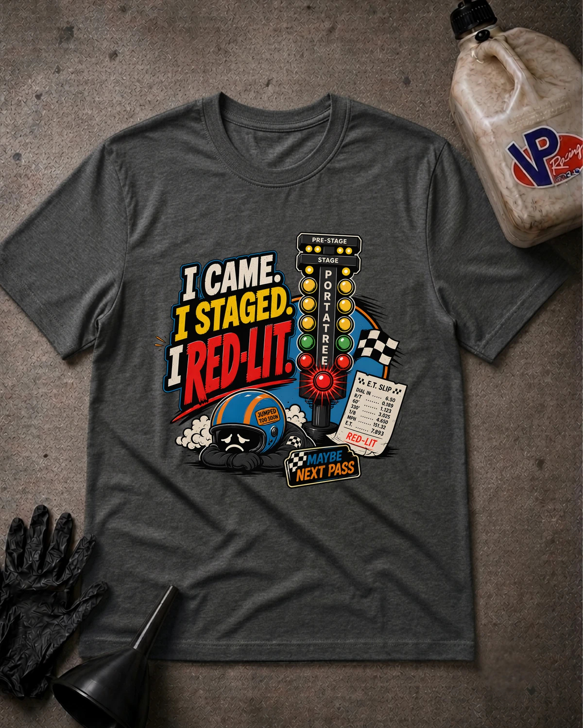 A dark heather gray graphic t-shirt laid flat on a textured gray surface, surrounded by racing-themed props including a black glove, funnel, and a plastic jug. The shirt features a colorful graphic with the text 'I CAME. I STAGED. I RED-LIT.'