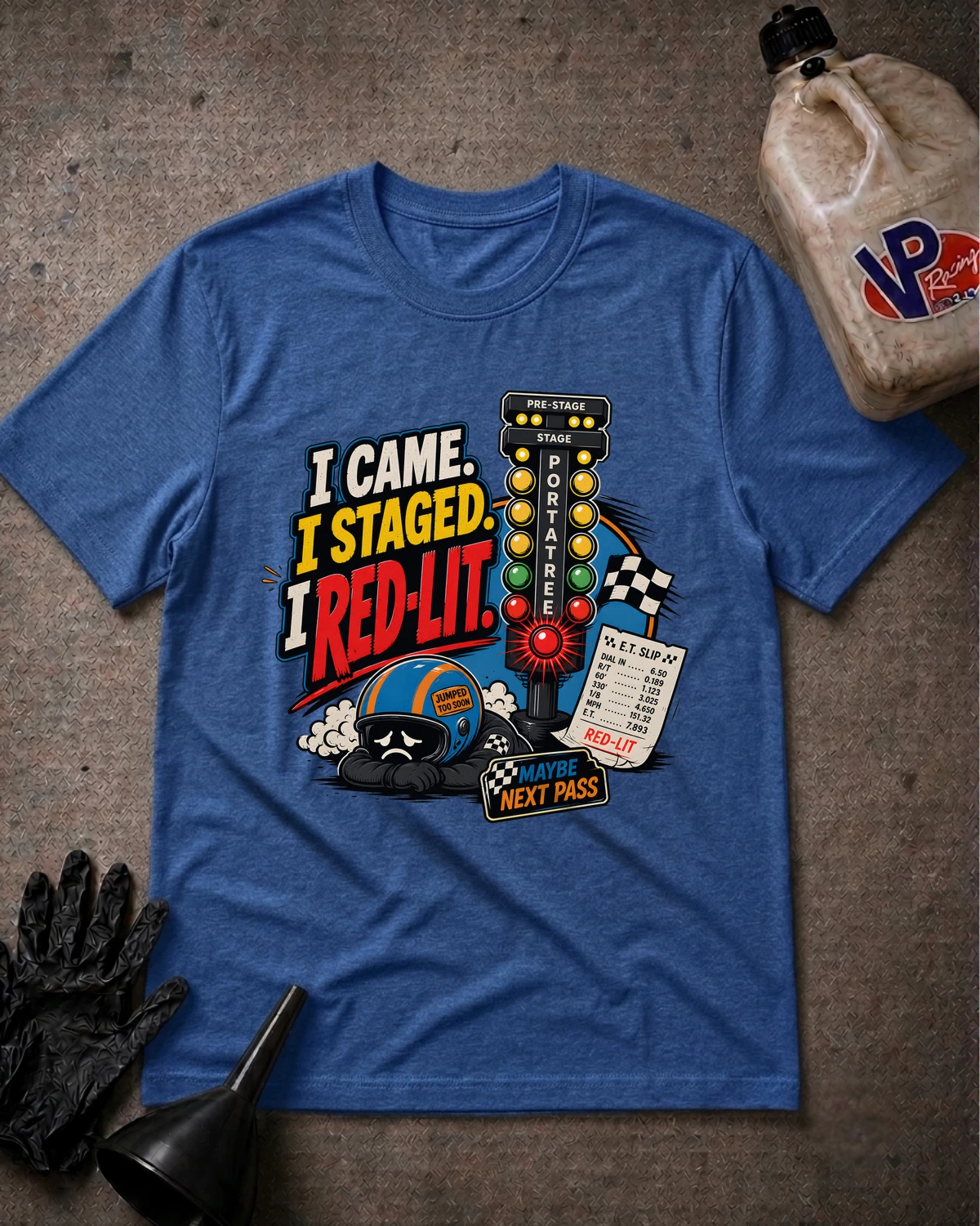 A heather blue graphic t-shirt laid flat on a textured gray surface, surrounded by racing-themed props including a black glove, funnel, and a plastic jug. The shirt features a colorful graphic with the text 'I CAME. I STAGED. I RED-LIT.'