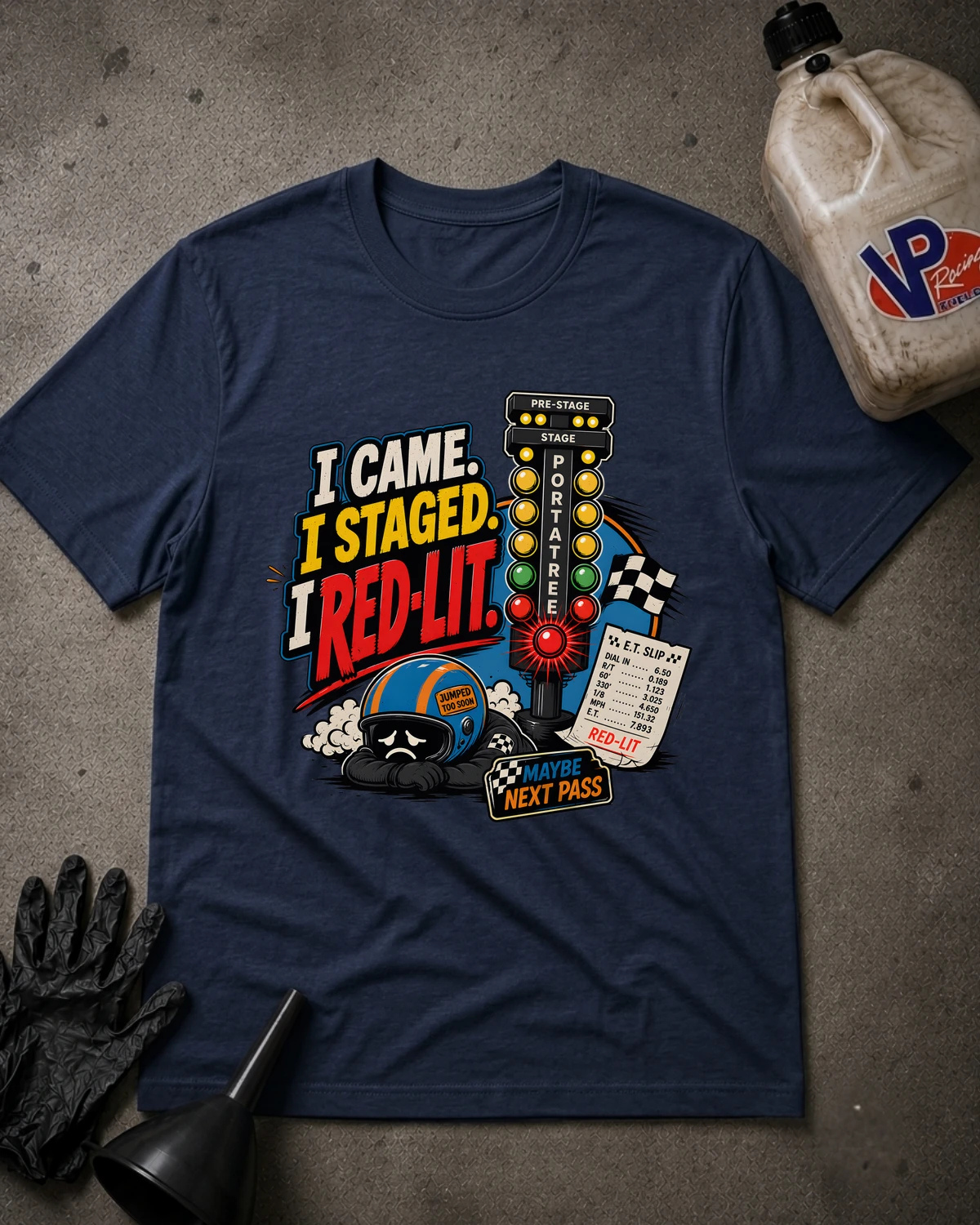 A navy blue graphic t-shirt laid flat on a textured gray surface, surrounded by racing-themed props including a bottle, gloves, and funnel. The shirt features a colorful graphic with the text 'I CAME. I STAGED. I RED-LIT.'