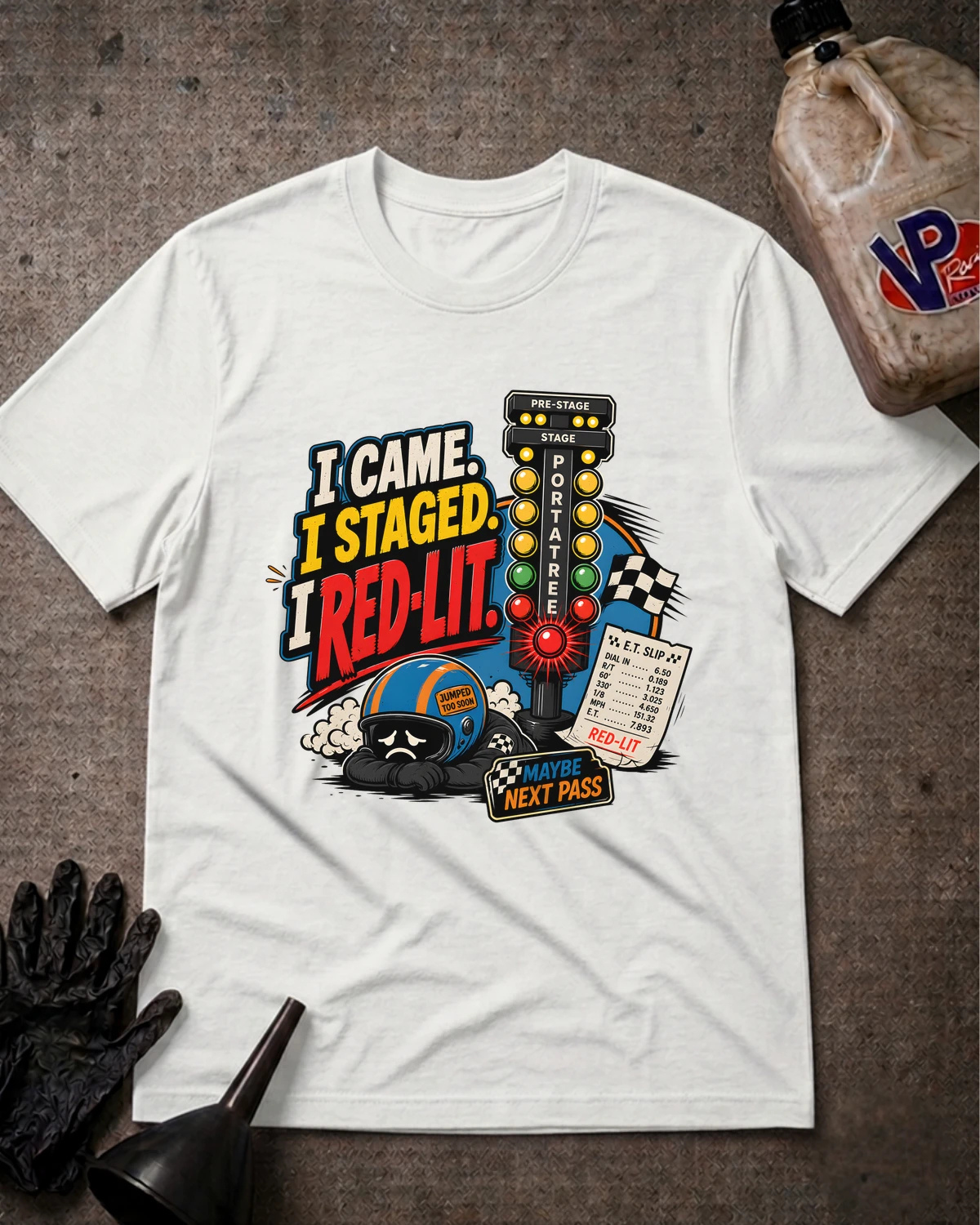 A white graphic t-shirt laid flat on a dark textured surface, featuring a colorful race track graphic with the text 'I CAME, I STAGED, I RED-LIT' and a race car, helmet, checkered flag, and pit pass. A black oil can, black gloves, and a dirty plastic jug are styled around the shirt.