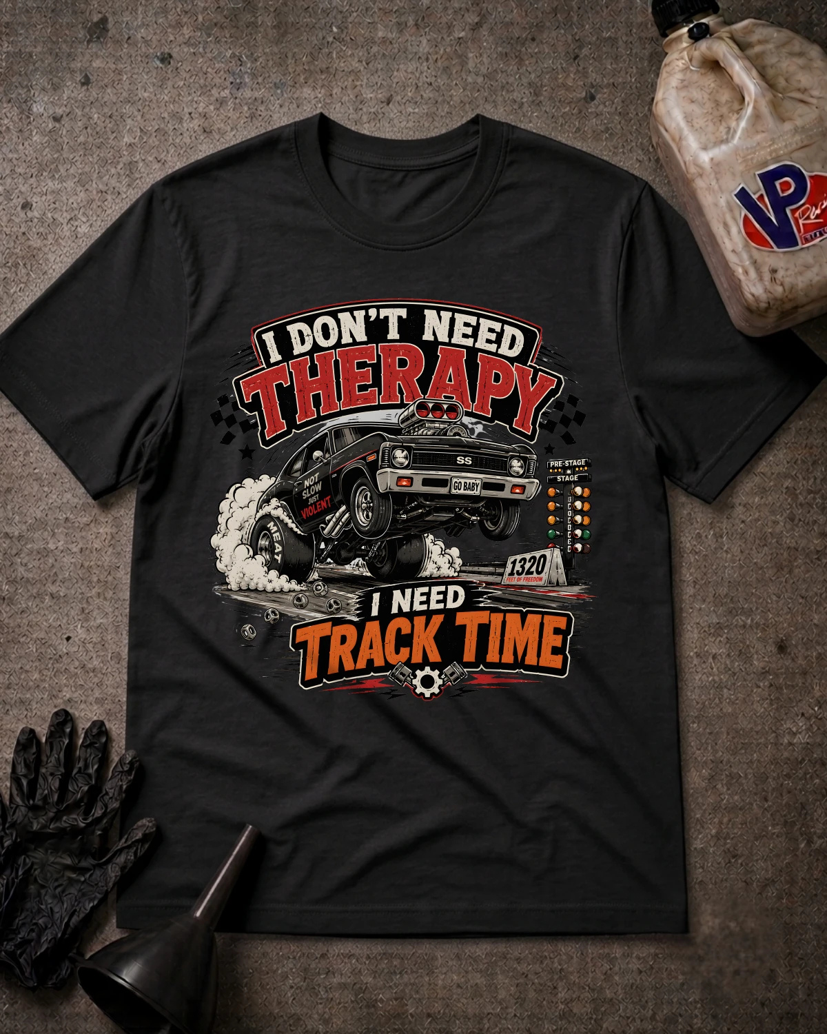 A black graphic t-shirt laid flat on a textured gray surface, surrounded by racing-themed props including a plastic jug, gloves, and a funnel. The shirt features a graphic of a black muscle car drifting with smoke, and the text 'I DON'T NEED THERAPY' and 'I NEED TRACK TIME' in red, white, and orange lettering.