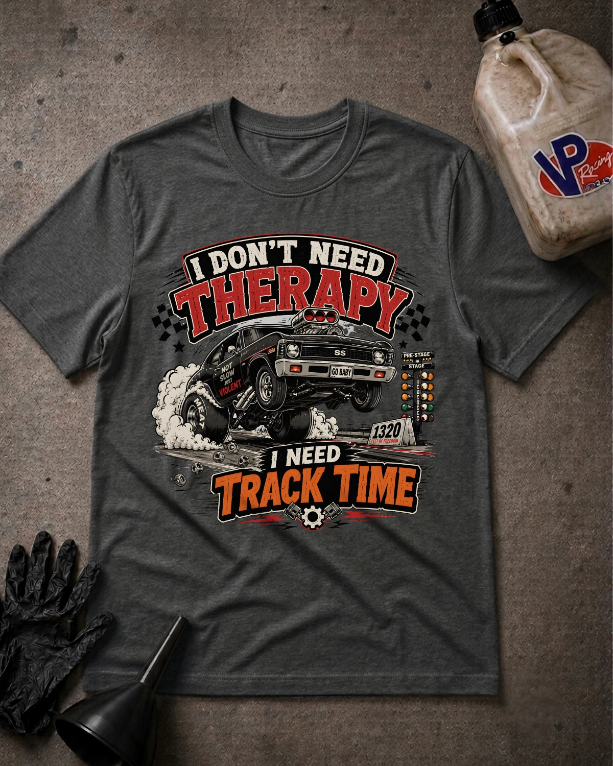 A dark heather gray graphic t-shirt laid flat on a textured gray surface, featuring a vintage-style car illustration and the text 'I DON'T NEED THERAPY I NEED TRACK TIME.' A black glove, funnel, and a plastic jug are placed around it as styled props.