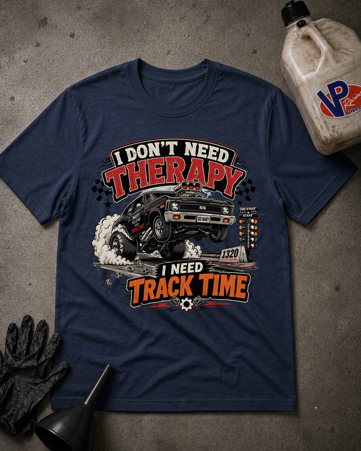 A navy blue graphic t-shirt laid flat on a gray concrete-like surface, surrounded by garage-themed props including black gloves, a funnel, and a plastic jug. The shirt features a large front print with the text 'I DON'T NEED THERAPY' and 'I NEED TRACK TIME' around an illustration of a black drag racing car kicking up smoke.