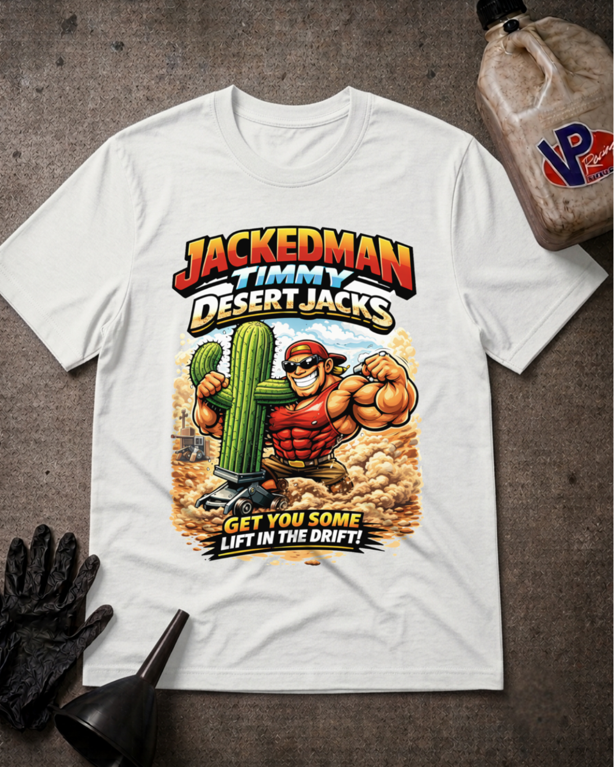 A white graphic t-shirt featuring a cartoon illustration of 'Jackedman Timmy Desert Jacks' flexing with a cactus. The shirt is displayed in a flat-lay arrangement on a dark textured surface, surrounded by black racing gloves, a black funnel, and a plastic bottle of oil.
