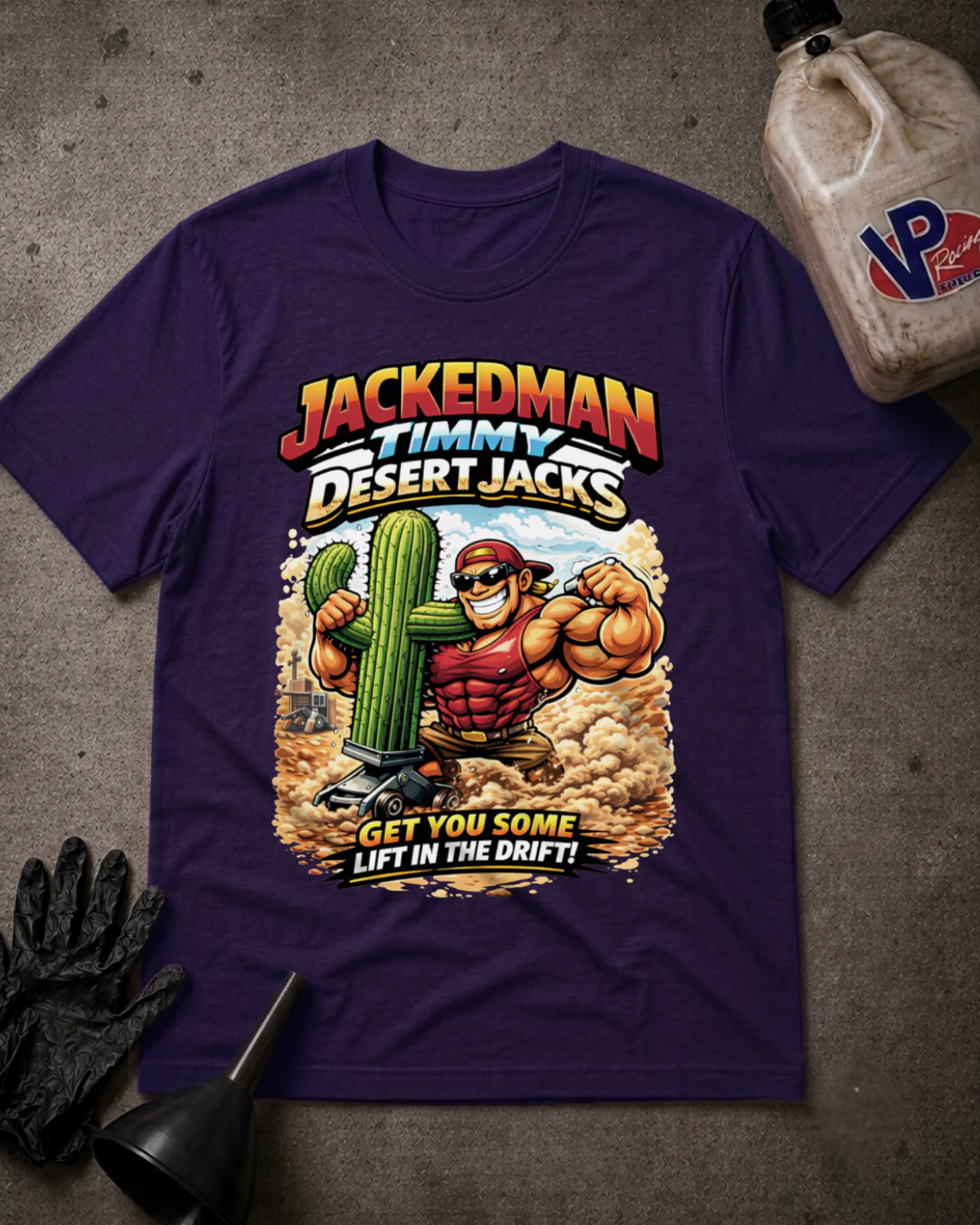 A purple short-sleeve graphic t-shirt laid flat on a textured gray surface, featuring a desert-themed cartoon illustration of a muscular man flexing next to a cactus with the text 'JACKEDMAN TIMMY DESERT JACKS' and 'GET YOU SOME LIFT IN THE DRIFT!'. A black glove, funnel, and a plastic jug are placed around it as props.