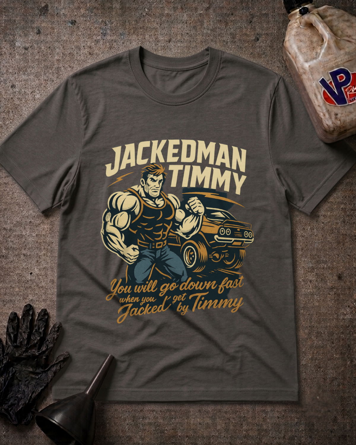 A dark gray graphic t-shirt featuring a muscular man flexing next to a muscle car with the text 'JACKEDMAN TIMMY' and 'You will go down fast when you get Jacked by Timmy'. The t-shirt is displayed in a flat-lay arrangement on a textured gray background with a black oil pan, black gloves, and a plastic jug.