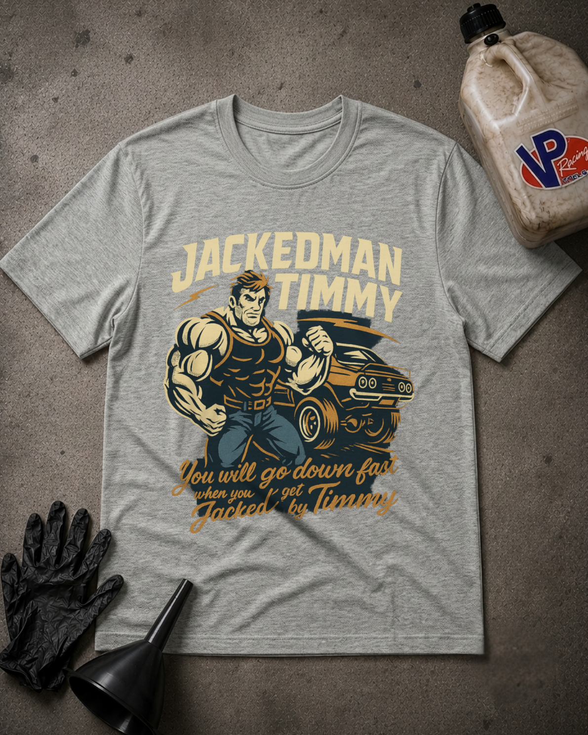 A heather gray graphic t-shirt featuring a muscular man flexing next to a muscle car with the text 'JACKEDMAN TIMMY' and 'You will go down fast when you get Jacked by Timmy'. The t-shirt is styled in a flat-lay on a gray concrete floor with black mechanic gloves, a funnel, and a plastic jug of VP Performance.
