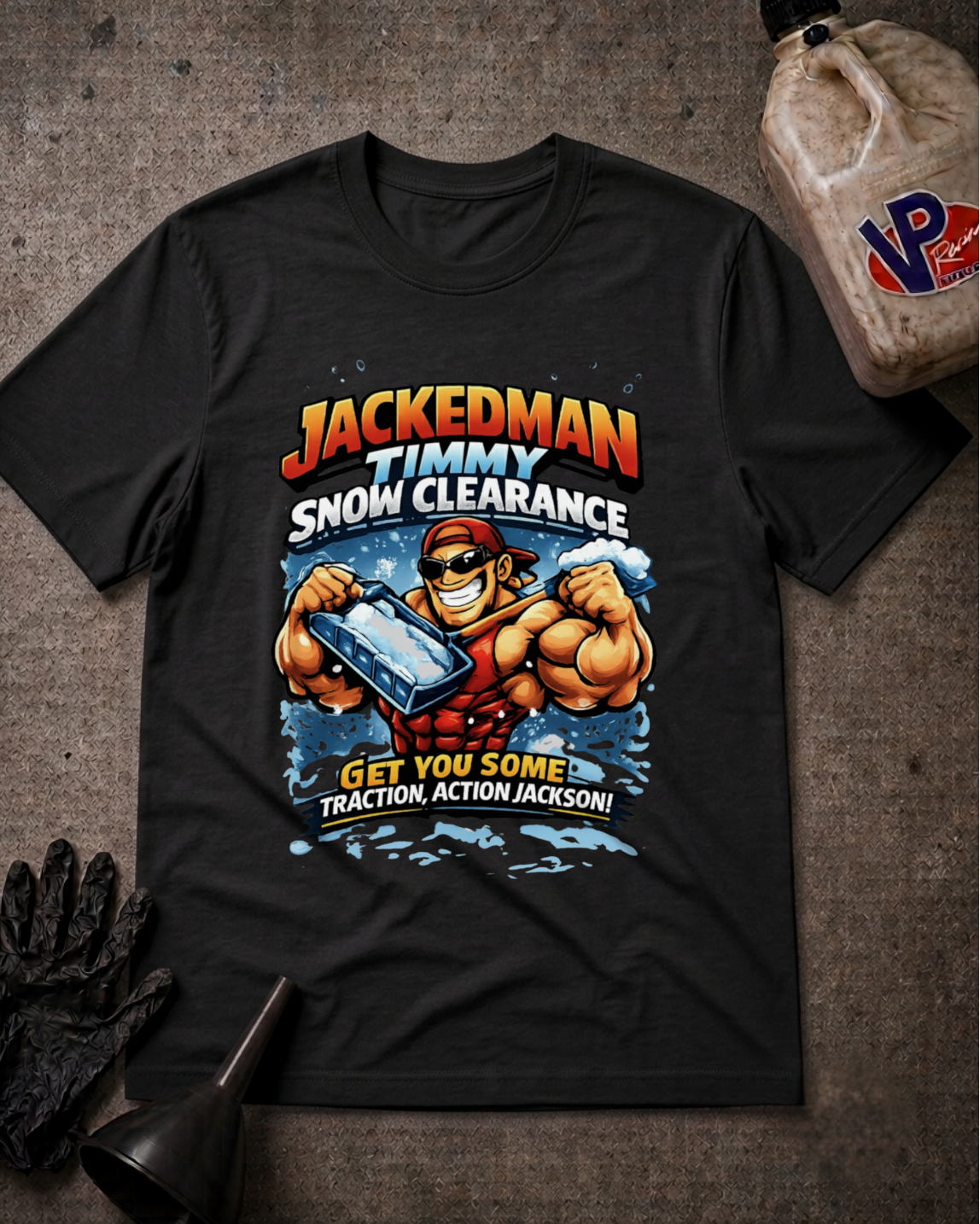 A black graphic t-shirt featuring a cartoon illustration of a muscular man in a red vest and hat clearing snow, with the text 'JACKEDMAN TIMMY SNOW CLEARANCE' and 'GET YOU SOME Traction, ACTION, JACKSON!'. The t-shirt is laid flat on a textured dark gray surface with a black glove, a funnel, and a plastic jug in the surrounding styled flat-lay props.