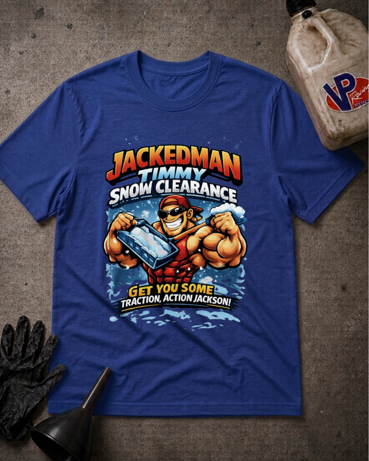 A royal blue graphic t-shirt featuring a cartoon illustration of a muscular man clearing snow with a chainsaw, with the text 'JACKEDMAN TIMMY SNOW CLEARANCE GET YOU SOME Traction, ACTION JACKSON!'. The t-shirt is displayed in a flat-lay arrangement on a textured gray concrete-like surface, accompanied by black work gloves, a black funnel, and a plastic jug in the corners.