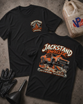 Two black graphic T-shirts laid flat on a textured gray surface, with automotive-themed artwork and the text 'JACKSTAND DWELLEER' on the front and back. The scene includes styling props such as a plastic jug, black gloves, and a funnel.