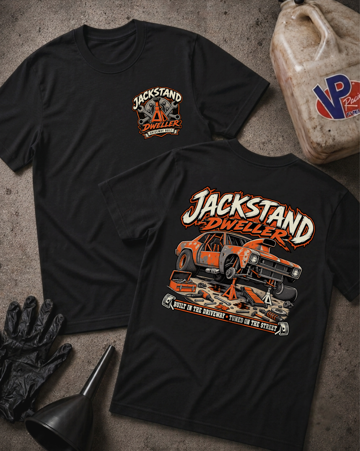 Two black graphic T-shirts laid flat on a textured gray surface, with automotive-themed artwork and the text 'JACKSTAND DWELLEER' on the front and back. The scene includes styling props such as a plastic jug, black gloves, and a funnel.