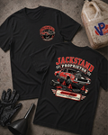 Two black graphic T-shirts laid flat on a gray concrete surface, surrounded by garage props including a VP Racing jug, black mechanic gloves, and a funnel. The front shirt features a circular left-chest graphic reading "Jackstand Proprietor," while the back shirt displays a large graphic of a vintage car up on jackstands with the text "Jackstand Proprietor" above and "Current Status: 3–5 Business Days From A Breakdown" below.