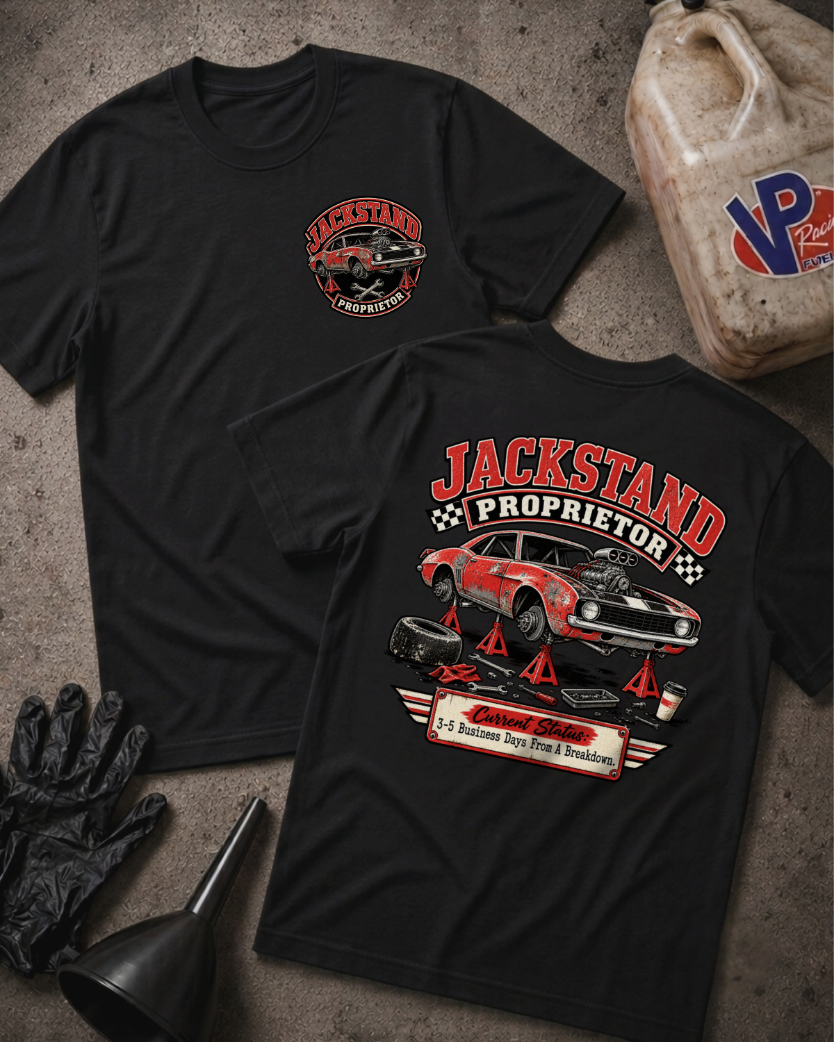 Two black graphic T-shirts laid flat on a gray concrete surface, surrounded by garage props including a VP Racing jug, black mechanic gloves, and a funnel. The front shirt features a circular left-chest graphic reading "Jackstand Proprietor," while the back shirt displays a large graphic of a vintage car up on jackstands with the text "Jackstand Proprietor" above and "Current Status: 3–5 Business Days From A Breakdown" below.