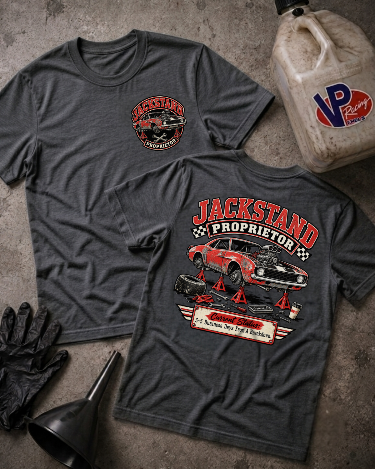 Two dark grey heather graphic T-shirts laid flat on a gray concrete surface, surrounded by garage props including a VP Racing jug, black mechanic gloves, and a funnel. The front shirt features a circular left-chest graphic reading "Jackstand Proprietor," while the back shirt displays a large graphic of a vintage car up on jackstands with the text "Jackstand Proprietor" above and "Current Status: 3–5 Business Days From A Breakdown" below.