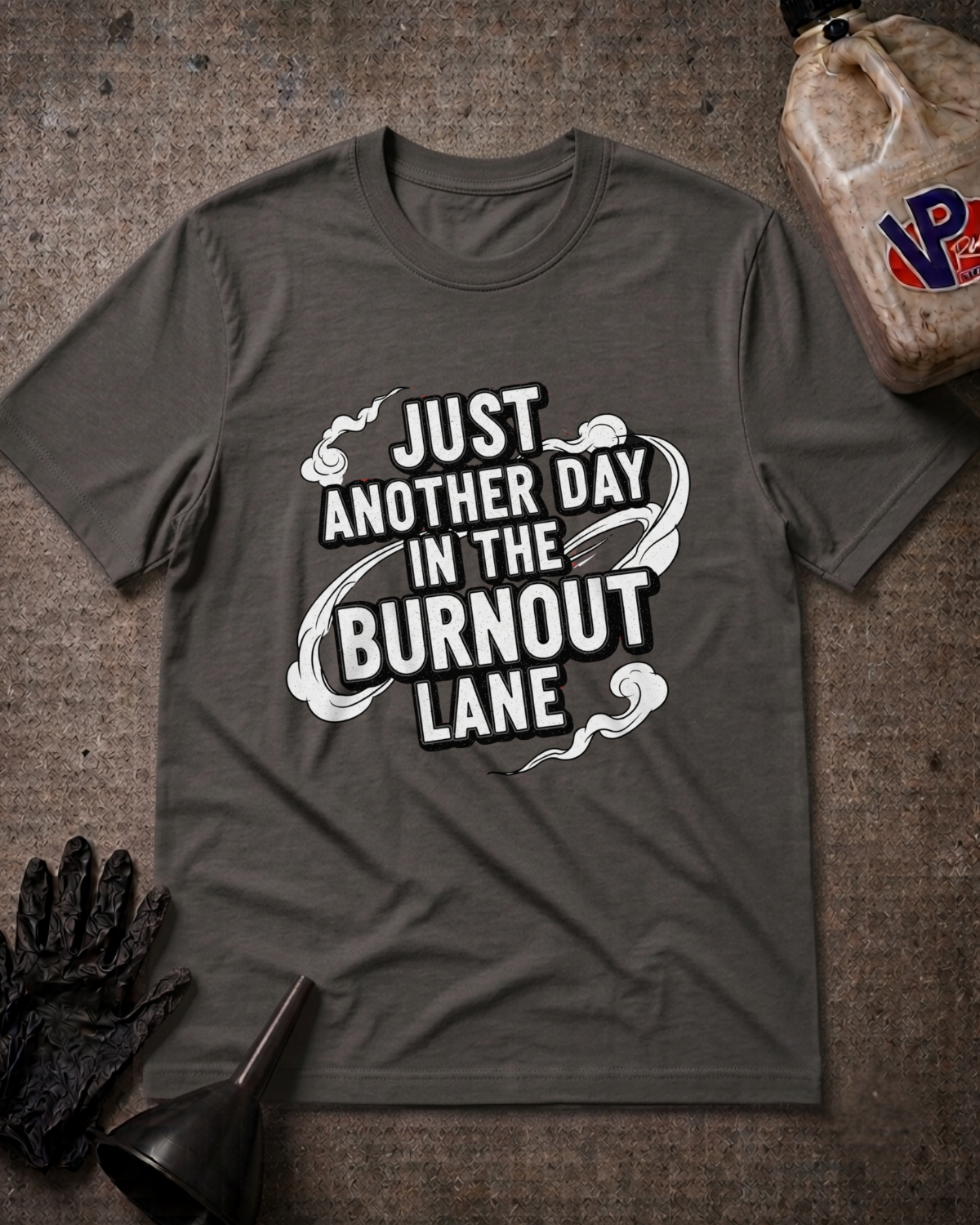 A dark gray crewneck t-shirt with a white graphic print reading 'JUST ANOTHER DAY IN THE BURNOUT LANE' styled in a flat-lay on a textured gray background with a black glove, funnel, and a fuel jug prop.