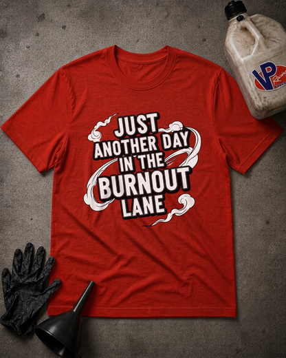 A red graphic t-shirt laid flat on a gray concrete-like surface, surrounded by burnout-themed props including a black funnel, black gloves, and a dirty plastic jug. The shirt features a bold white and black graphic that reads 'JUST ANOTHER DAY IN THE BURNOUT LANE'.