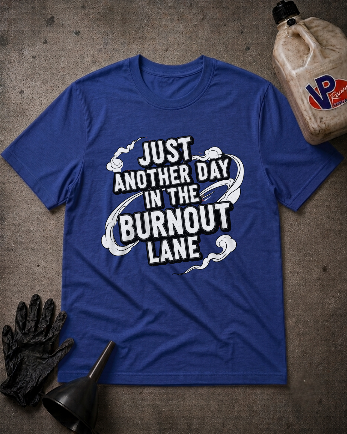 A royal blue graphic t-shirt laid flat on a textured gray surface, surrounded by automotive-themed props including a black glove, funnel, and a jug of VP gasoline. The t-shirt features a white graphic with the text 'JUST ANOTHER DAY IN THE BURNOUT LANE'.