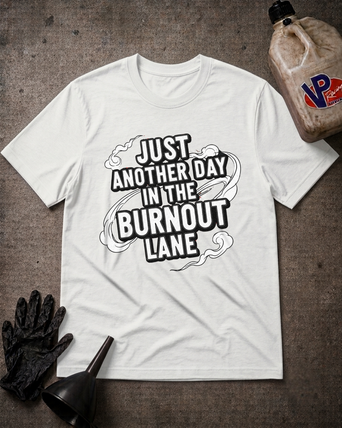 A white graphic t-shirt laid flat on a dark textured surface, surrounded by work-themed props including a black glove, funnel, and a jug of VP paint. The t-shirt features a bold black graphic that reads 'JUST ANOTHER DAY IN THE BURNOUT LANE'.