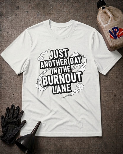 A white graphic t-shirt laid flat on a dark textured surface, surrounded by work-themed props including a black glove, funnel, and a jug of VP paint. The t-shirt features a bold black graphic that reads 'JUST ANOTHER DAY IN THE BURNOUT LANE'.