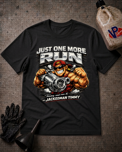 A black graphic t-shirt laid flat on a textured dark background, featuring a muscular cartoon character holding a large engine block with the text 'JUST ONE MORE RUN' and 'JACKEDMAN TIMMY'. A black glove, funnel, and a plastic jug are placed around it as styling props.