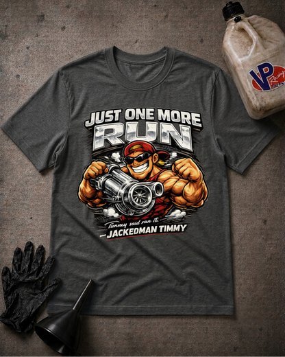 A dark heather gray graphic t-shirt laid flat on a textured gray surface, surrounded by racing-themed props including a black glove, a funnel, and a plastic fuel jug. The t-shirt features a large graphic of a muscular cartoon character holding a megaphone with the text 'JUST ONE MORE RUN' and 'JACKEDMAN TIMMY'.