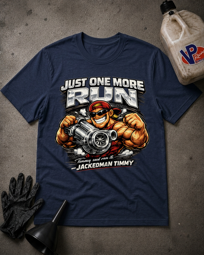A navy blue graphic t-shirt laid flat on a textured gray surface, surrounded by racing-themed props including a black glove, funnel, and a plastic jug. The t-shirt features a large graphic of a muscular race car driver with the text 'JUST ONE MORE RUN' and 'JACKEDMAN TIMMY'.