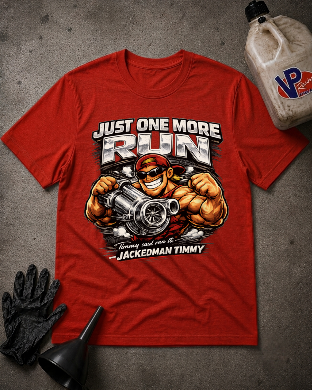 A red graphic t-shirt featuring a muscular cartoon character holding a large engine block, with the text 'JUST ONE MORE RUN' and 'JACKEDMAN TIMMY'. The shirt is displayed in a flat-lay arrangement on a gray concrete-like surface with black gloves, a funnel, and a plastic jug nearby.
