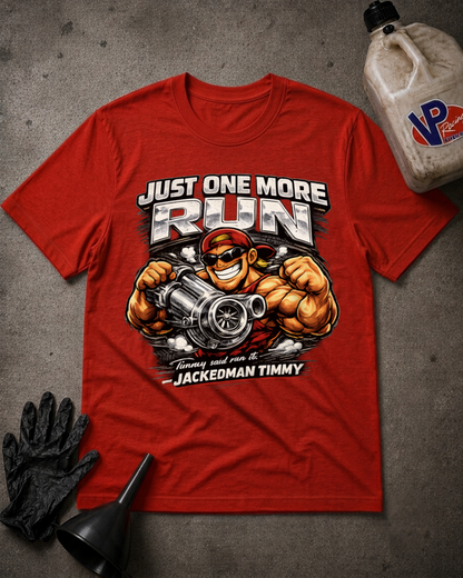 A red graphic t-shirt featuring a muscular cartoon character holding a large engine block, with the text 'JUST ONE MORE RUN' and 'JACKEDMAN TIMMY'. The shirt is displayed in a flat-lay arrangement on a gray concrete-like surface with black gloves, a funnel, and a plastic jug nearby.