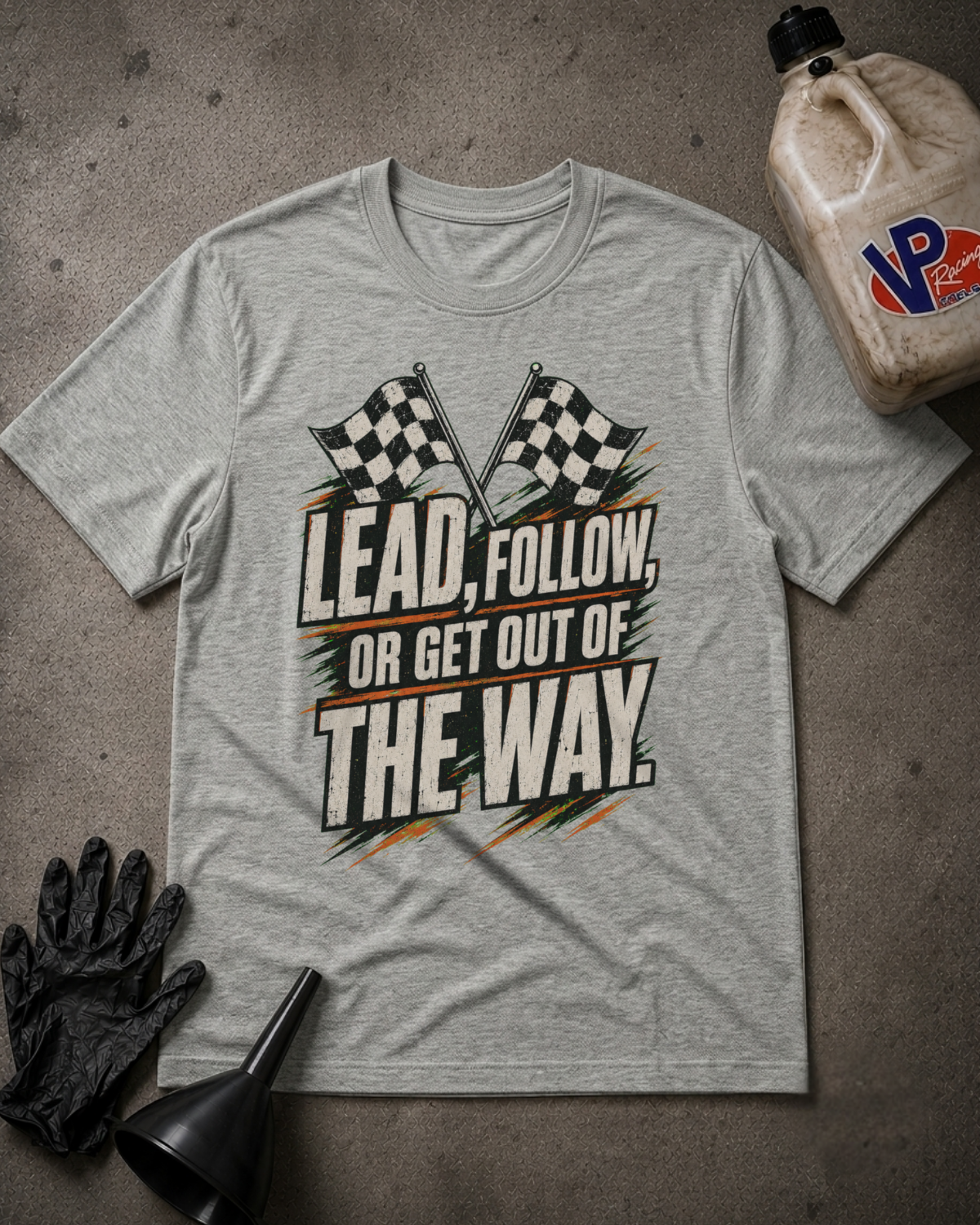 A light heather gray graphic t-shirt laid flat on a textured gray surface, surrounded by racing-themed props including a black oil bottle, a black funnel, black gloves, and a plastic jug. The t-shirt features a graphic with two checkered flags and the text 'LEAD, FOLLOW, OR GET OUT OF THE WAY.