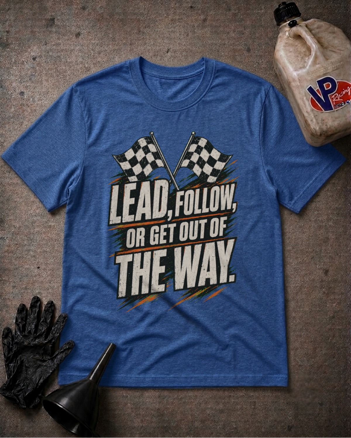 A heather blue graphic t-shirt laid flat on a textured gray surface, surrounded by racing-themed props including a black glove, a black funnel, and a plastic jug of VP Racing oil. The t-shirt features a graphic with two checkered flags and the text 'LEAD, FOLLOW, OR GET OUT OF THE WAY.