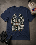 A navy blue graphic t-shirt laid flat on a gray concrete surface, surrounded by racing-themed props including a black glove, a black funnel, and a jug of VP Racing oil. The t-shirt features a graphic with checkered flags and the text 'LEAD, FOLLOW, OR GET OUT OF THE WAY.
