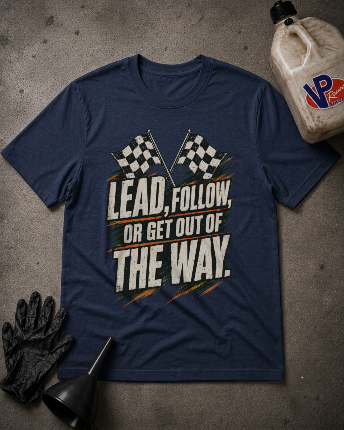 A navy blue graphic t-shirt laid flat on a gray concrete surface, surrounded by racing-themed props including a black glove, a black funnel, and a jug of VP Racing oil. The t-shirt features a graphic with checkered flags and the text 'LEAD, FOLLOW, OR GET OUT OF THE WAY.