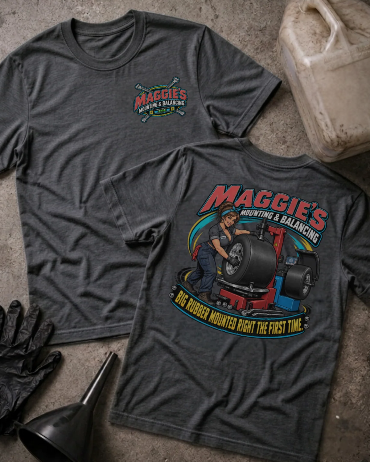 Front and back of Maggie’s Mounting & Balancing dark grey heather T-shirt with chest logo, woman working with tire equipment, and “Big rubber mounted right the first time” slogan.
